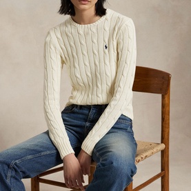 Model wearing Polo Ralph Lauren Authentic Cream cable-knit sweater with navy pony logo, styled with blue jeans, seated on a wooden chair.