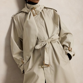 Model wearing Polo Ralph Lauren Buckled Scarf Hooded Trench Coat in Classic Stone with self-belt, brass buckle, and belted neck scarf detail.