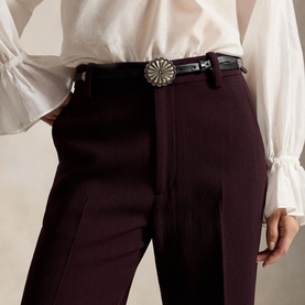 Model wearing Polo Ralph Lauren Midnight Merlot trousers with a black leather concho belt and white ruffled blouse.