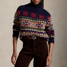 Model wearing navy Fair Isle turtleneck jumper with red, yellow and purple snowflake pattern, styled with brown cord trousers and belt
