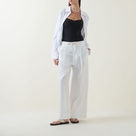 Model wearing Sea Level black swimsuit with white wide-leg trousers, white shirt and brown flat sandals, pearl necklace