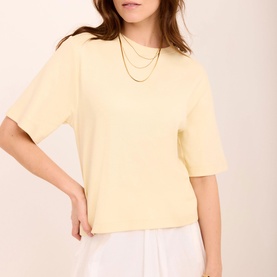 Model wearing Mint Velvet yellow boxy short-sleeve top, styled with white trousers and layered gold chain necklaces.