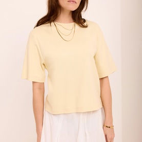 Model wearing Mint Velvet yellow boxy short-sleeve top, styled with layered gold necklaces and white wide-leg trousers.