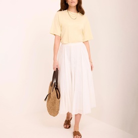 Model wearing Mint Velvet yellow short-sleeve top with white midi pleated skirt, styled with woven tote bag and tan sandals.
