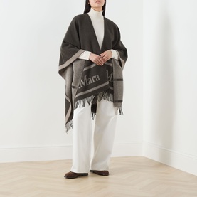 Model wearing Max Mara Accessories Hilde Logo Fringed Cape in dark brown with contrast border and bold logo, styled with white wide-leg trousers