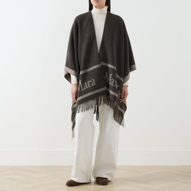 Model wearing Max Mara Accessories Hilde Logo Fringed Cape in dark brown with bold logo detail and fringed hem, styled with white wide-leg trousers