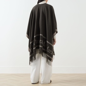 Model wearing Max Mara Accessories Hilde Logo Fringed Cape in dark brown with bold logo detail and tassel hem, styled with white wide-leg trousers.