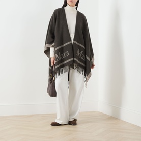 Model wearing Max Mara Accessories Hilde Logo Fringed Cape in dark brown with bold logo, fringed hem, styled with white wide-leg trousers.