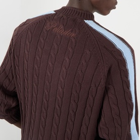 Model wearing Pellador Overlap Cable Knit Sweater in brown with light blue double-stripe sleeve detail and embroidered logo at chest.