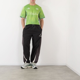 Model wearing Pellador Logo Football T-Shirt in green jersey style, paired with black track pants and grey trainers.