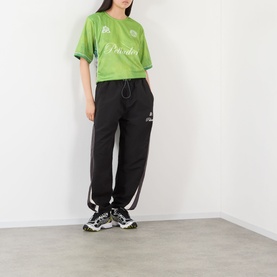 Model wearing Pellador Logo Football T-Shirt in green jersey style with black logo, styled with black joggers and chunky trainers.