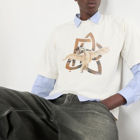Model wearing Pellador Swan Logo T-Shirt in white with front graphic, layered over blue shirt, styled with dark grey jeans.