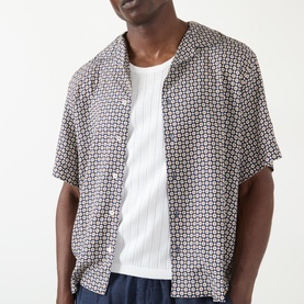 Model wearing A.P.C. Bennet Printed Shirt in dark navy geometric print, camp collar, short sleeves, styled with white ribbed vest and navy trousers.