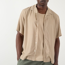 Model wearing A.P.C. beige geometric print short-sleeve camp collar shirt in viscose, styled over a plain beige tee with khaki trousers.