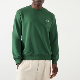 Model wearing A.P.C. dark green cotton sweatshirt with small white logo print at chest, styled with cream trousers.