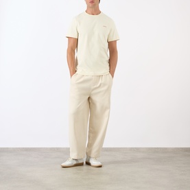 Model wearing A.P.C. Oliver Logo T-Shirt in white, round neck, short sleeves, styled with matching cream wide-leg trousers and white trainers.