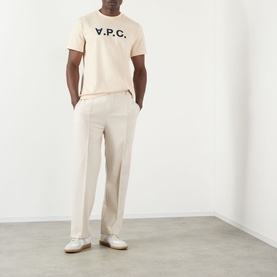 Model wearing A.P.C. VPC T-shirt in mastic with navy logo, styled with cream wide-leg trousers and white trainers.