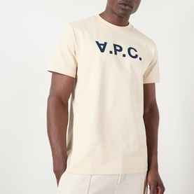 Model wearing A.P.C. T-Shirt in mastic with navy V.P.C. logo print, crew neck, styled with light grey trousers.