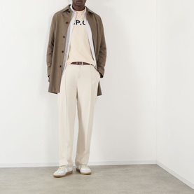 Model wearing A.P.C. logo T-shirt in mastic, styled with brown overcoat, cream trousers, brown belt and white trainers.