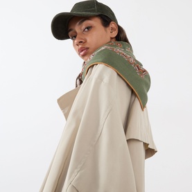 Model wearing beige oversized coat with khaki paisley scarf and matching green cap, shot from behind at an angle