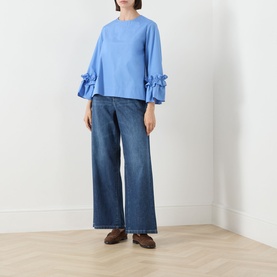 Model wearing Max Mara S light blue cotton shirt with ruffle-trimmed sleeves, styled with wide-leg blue jeans and brown loafers.