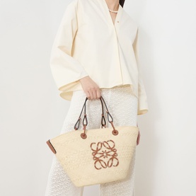 Model carrying Loewe Anagram Medium Basket Bag in natural, woven iraca palm with tan leather handles, embroidered anagram logo detail.
