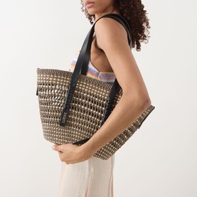 Model carrying Loewe Braid Medium Braid Basket Bag in khaki/natural woven iraca with black leather handles, styled with cream trousers and printed