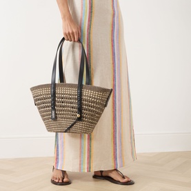 Model carrying Loewe Braid Medium Braid Basket Bag in khaki/natural woven iraca with dark leather handles, styled with striped maxi dress and sandals