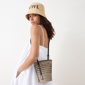 Model carrying Loewe Braid Pocket Crossbody Bag in khaki/natural, woven iraca with black leather strap. Styled with white dress and Loewe bucket hat.