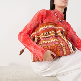 Model carrying Loewe Flamenco Crochet Medium Clutch in bright orange and pink raffia with ruffled top, styled with red lace top and white trousers