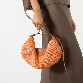 Model carrying Loewe Swing Medium Shoulder Bag in bright orange and pink woven raffia with tan leather strap and fortune cookie shape