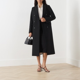 Model wearing Max Mara Studio black skirt, styled with a long black coat, white shirt, and strappy heeled sandals, carrying a black handbag.