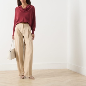 Model wearing Max Mara Studio ruby draped cowl-neck top, styled with wide-leg beige trousers and carrying a white handbag.