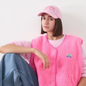 Model with Ganni Logo Washed Baseball Cap in Pink Nectar. Styled with pink sherpa vest and blue jeans.