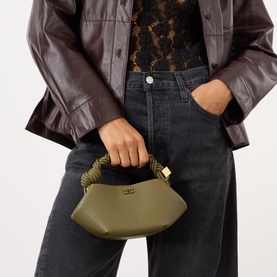 Model carrying Ganni Bou Mini Crossbody Bag in Military Olive, braided top handle, gold-tone hardware, styled with dark jeans and burgundy leather