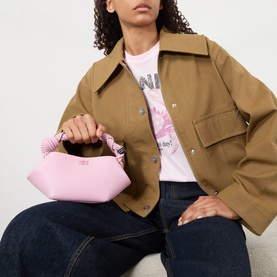 Model carrying Ganni Bou Mini Crossbody Bag in Pink Nectar with braided top handle and silver-tone hardware, styled with brown jacket and jeans.