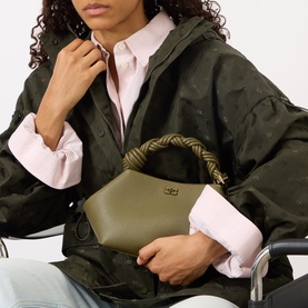 Model carrying Ganni Bou Small Crossbody Bag in Military Olive with braided top handle and gold-tone hardware, styled with khaki jacket and pink