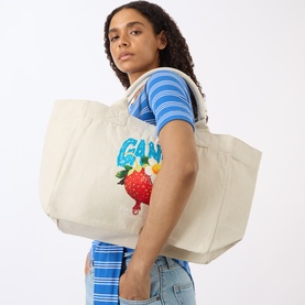 Model carrying Ganni Strawberry Logo Medium Tote Bag in Sunlight canvas with strawberry graphic print, styled with blue striped tee and jeans.