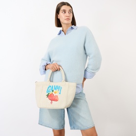 Model carrying Ganni Strawberry Logo Small Tote Bag in Sunlight canvas with strawberry graphic print, styled with blue knit and denim shorts