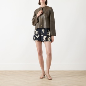 Model wearing Max Mara Main Bouquet Cropped Leather Jacket in kaki, collarless with button fastening, styled with printed shorts and heeled sandals.