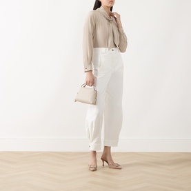 Model wearing Max Mara Dry Bow-Neck Silk Blouse in stone, styled with white wide-leg trousers, beige heels, and a small top-handle bag.