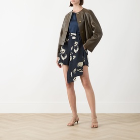Model wearing a navy abstract-print mini dress with asymmetric hem, styled with an olive green collarless jacket and nude heeled mules.