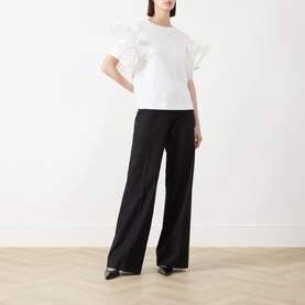 Model wearing Max Mara Main Costone Ruffle T-Shirt in white with taffeta ruffle sleeves, styled with wide-leg black trousers and black heels.