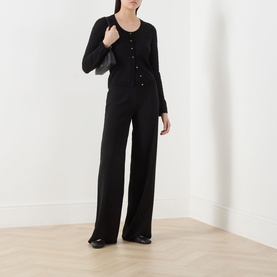 Model wearing black wide-leg trousers and black button-front cardigan, carrying a black shoulder bag, with black flat shoes