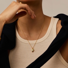 Model with Missoma gold necklace featuring a small letter pendant on a fine chain, styled with a cream ribbed vest and black jacket