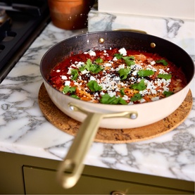 Stellar Cast Light Saut&eacute; Pan 28cm in clay with gold-tone handle, shown on marble surface with tomato and feta dish cooking inside.