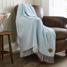 The Lyndon Company Arcadia Throw Blue draped over a brown leather armchair, with white fringe trim and woven texture.