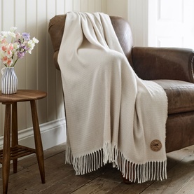 The Lyndon Company Arcadia Throw in Doeskin, draped over a brown leather armchair with fringe hem detail and branded leather patch.