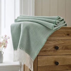 The Lyndon Company Arcadia Throw in sage green, textured knit weave with cream fringe trim, draped over a wooden chest of drawers
