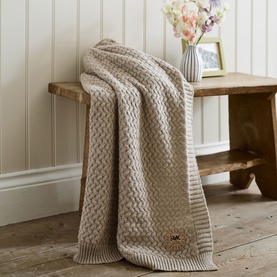 The Lyndon Company Cadenza Throw in Natural, cable-knit texture, draped over a rustic wooden bench in a cosy home setting.